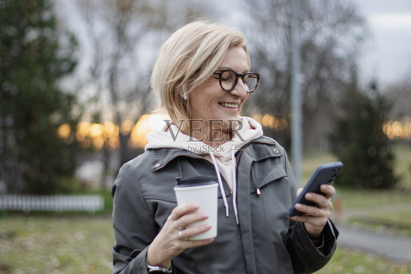 一位成熟的金发女性戴着眼镜,在城市公园里散步并享受着使用手机的时光。照片摄影图片