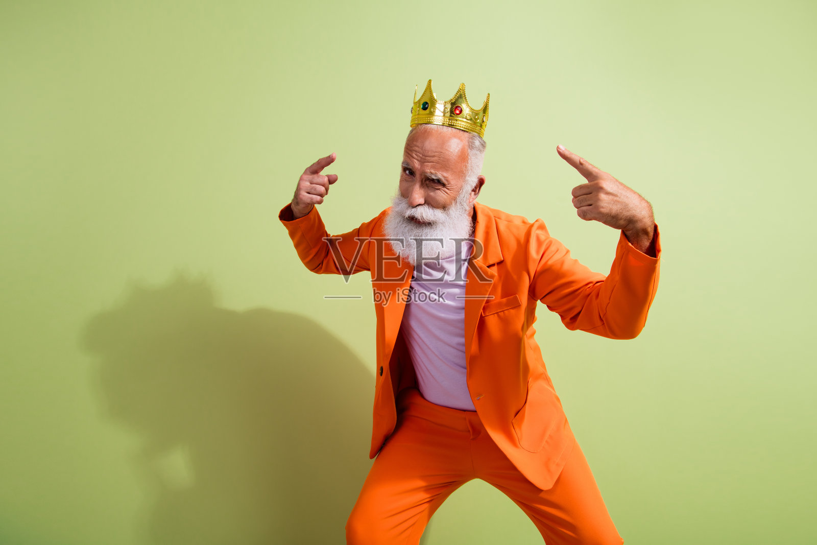 搞笑的表演者指着空白处,戴着金色王冠,穿着橙色西装,背景为绿色。照片摄影图片