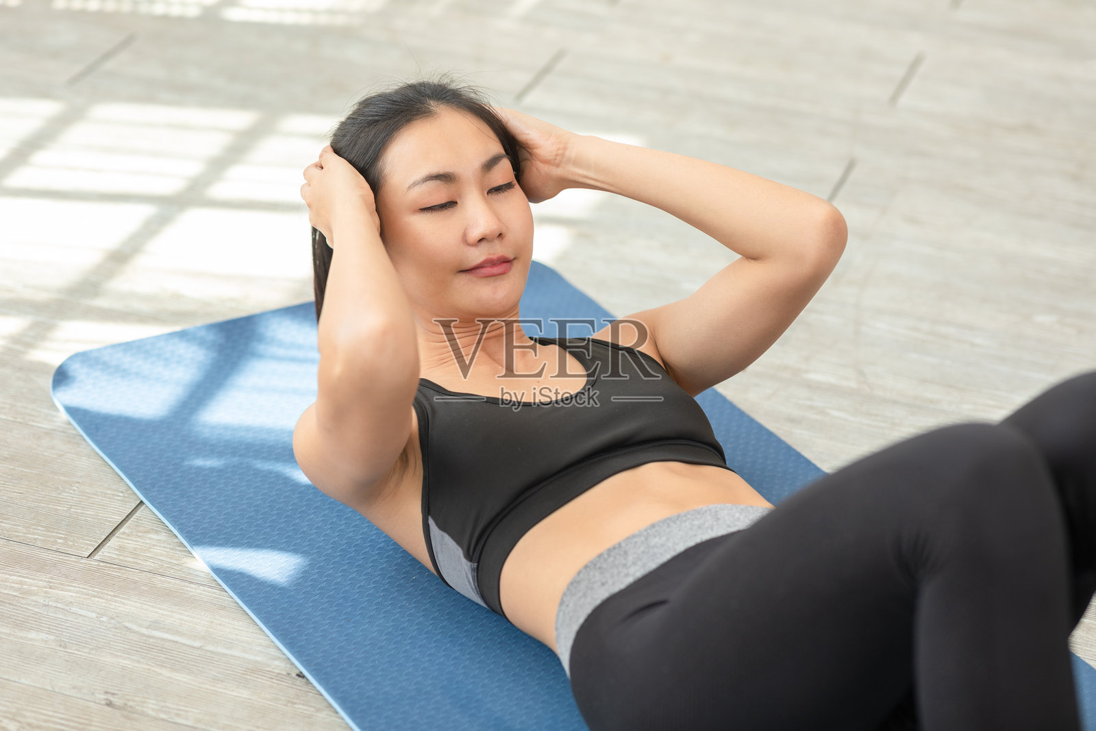 一位亚洲女性在明亮的家居环境中,穿着运动服,在蓝色健身垫上做仰卧起坐,展现出对健身和健康生活方式的坚持。照片摄影图片
