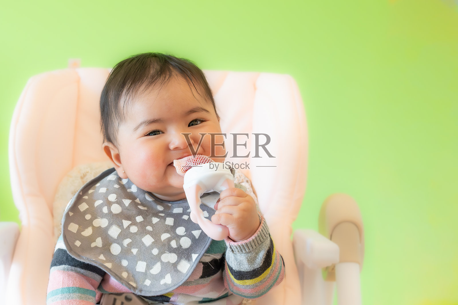 快乐的亚洲宝宝独自用自动喂养奶嘴吃水果。坐在粉色高脚椅上,背景为绿色,留有空白空间。为小朋友介绍新鲜食物的有趣健康方式。照片摄影图片