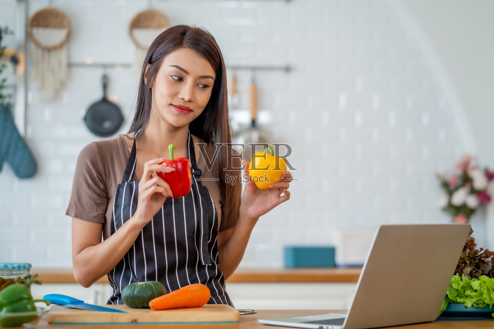 穿着围裙的女人手持红色和黄色的甜椒,专注地看着现代厨房里的笔记本电脑。她看起来专注且投入于烹饪。照片摄影图片