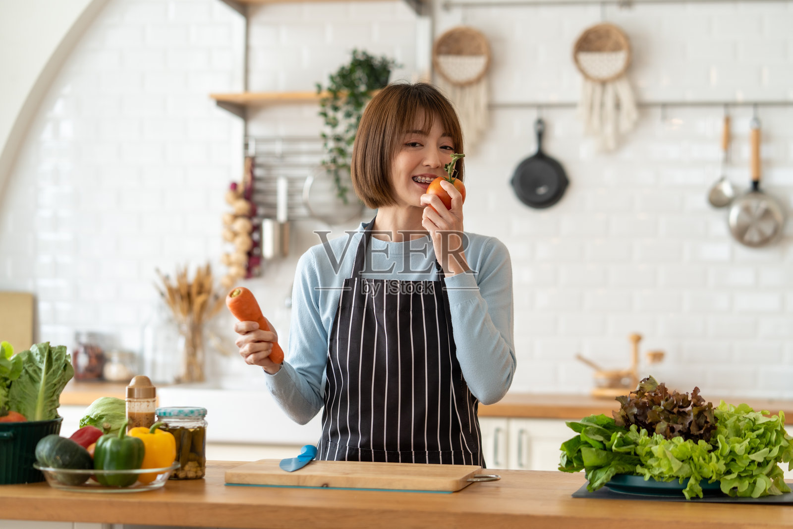 女人在厨房里手持胡萝卜和甜椒,正在准备新鲜蔬菜。她微笑着享受烹饪的乐趣。照片摄影图片