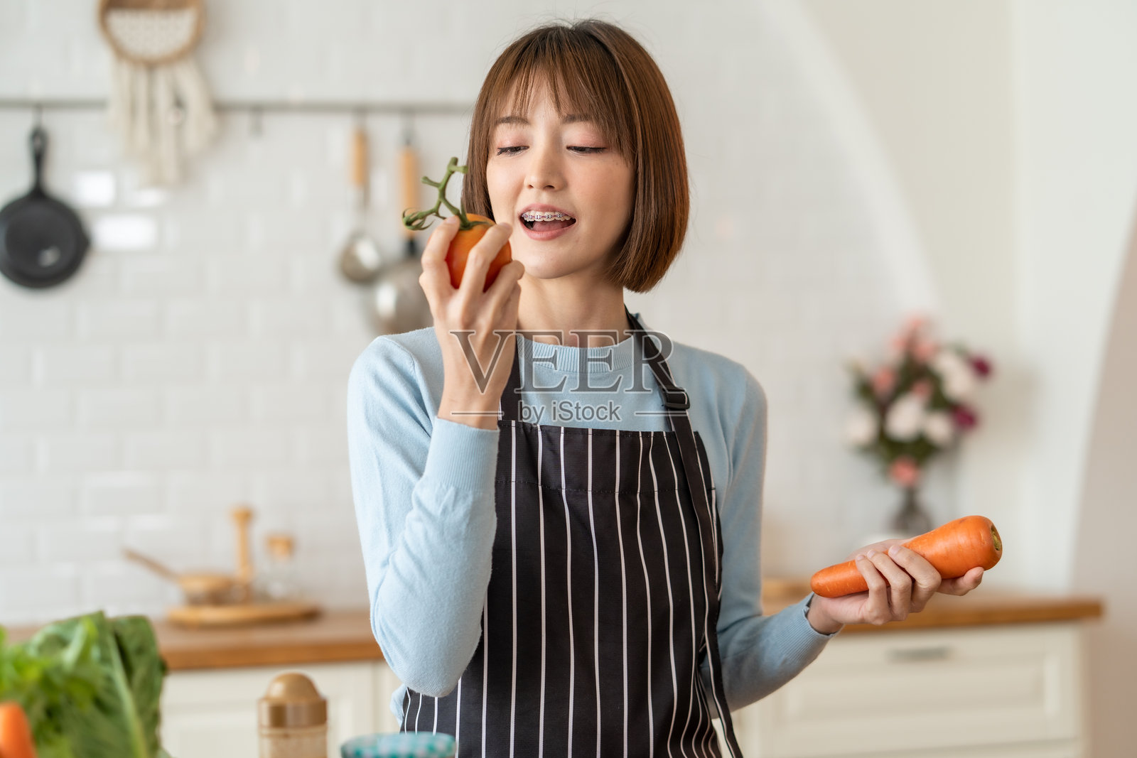 穿围裙的女人在明亮的厨房里检查番茄,同时手里拿着胡萝卜。她看起来专注而投入于烹饪,周围是新鲜的蔬菜。照片摄影图片