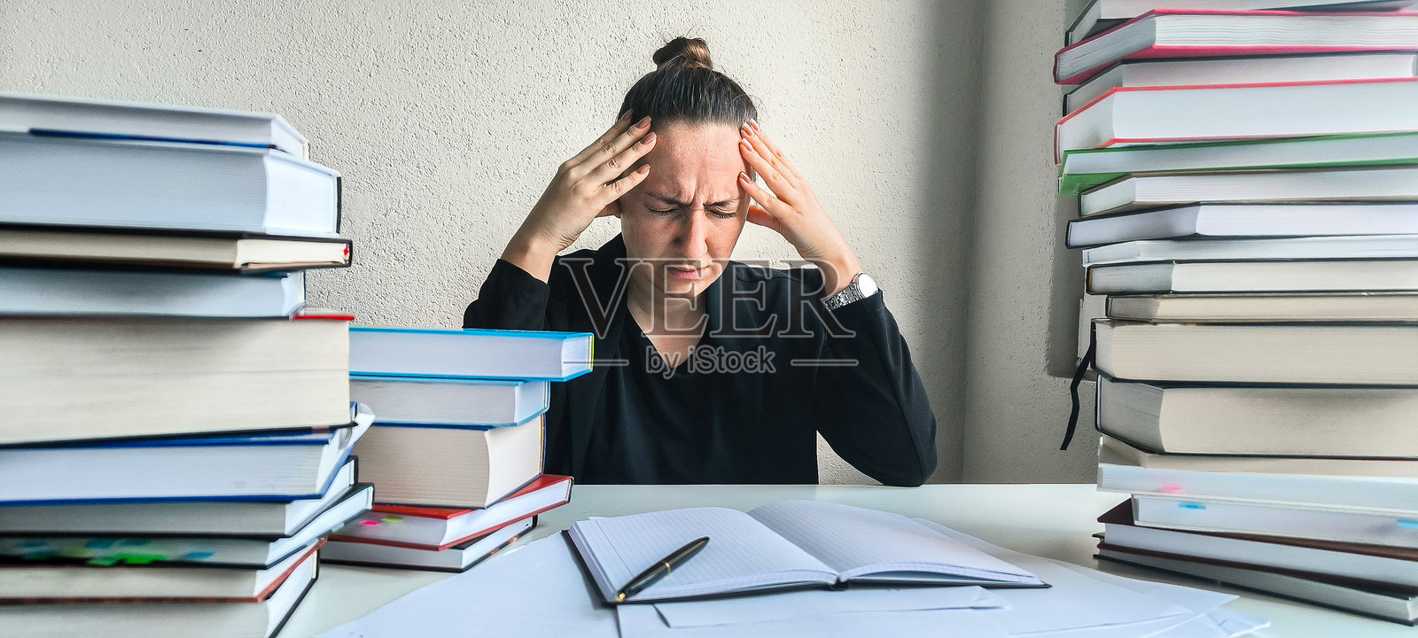 一位穿着商务套装的年轻女性坐在桌子前,桌上有文件和一堆书籍。工作和大学的压力。情感疲惫。违反工作与休息的时间安排。21世纪的问题。照片摄影图片