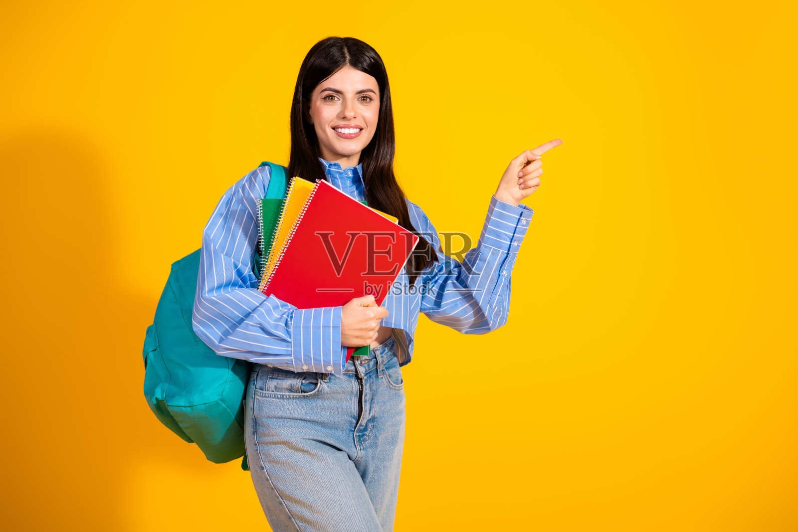 年轻女学生在鲜艳的黄色背景前摆姿势,穿着休闲,面带自信微笑,手中拿着书籍。照片摄影图片