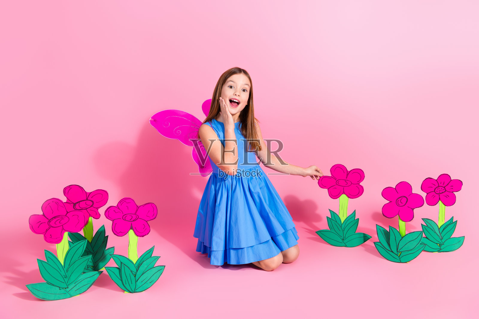 穿着蓝色裙子的快乐小女孩在粉色背景前摆姿势,周围环绕着五彩缤纷的花卉装饰。照片摄影图片