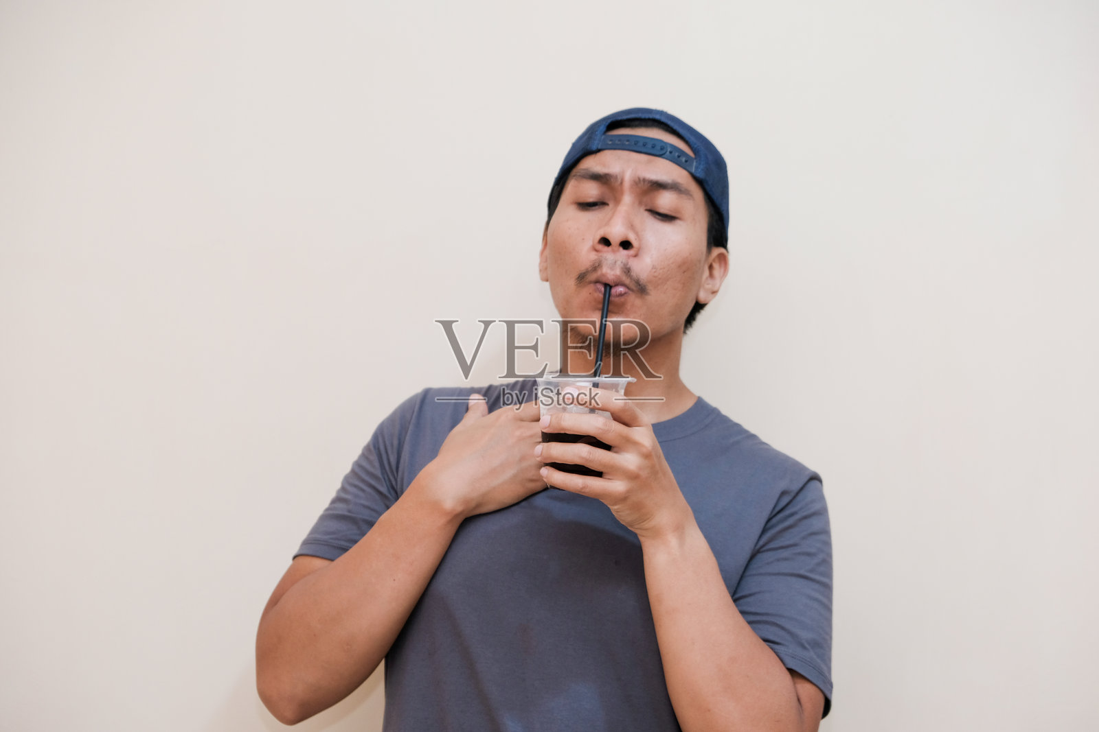 一位东南亚男子穿着休闲服,手持可乐饮料,背景为纯色。照片摄影图片