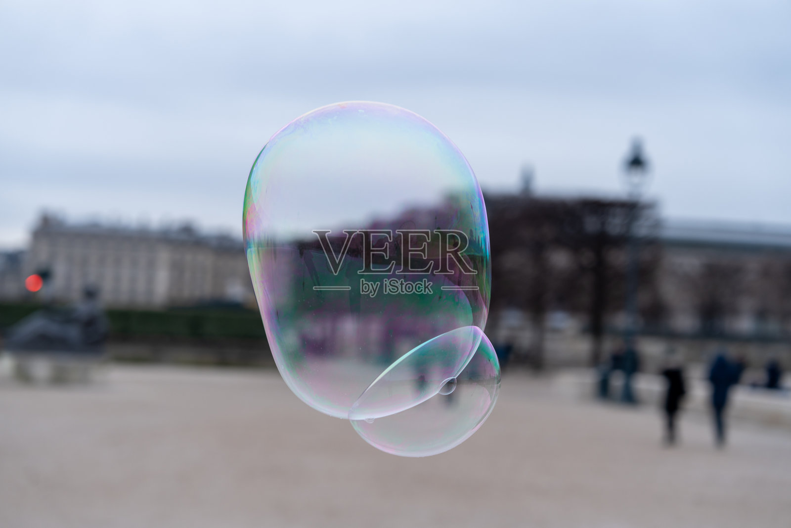 五彩斑斓的肥皂泡在历史公园中漂浮,背景中有行人悠闲地散步,天气阴云密布。照片摄影图片