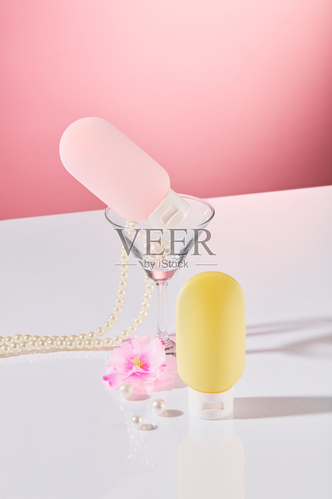 粉色背景上,两款瓶子的特写,颜色为粉色和黄色,还有一只马提尼酒杯、杏花和一串珍珠线。这是一则以派对为主题的化妆品广告。照片摄影图片