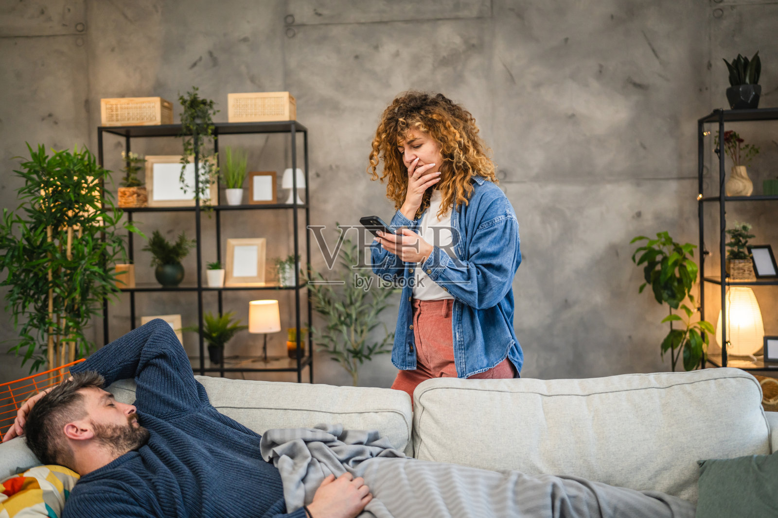 震惊的女人在沙发上工作的同时使用手机,而男人则在沙发上睡觉。照片摄影图片