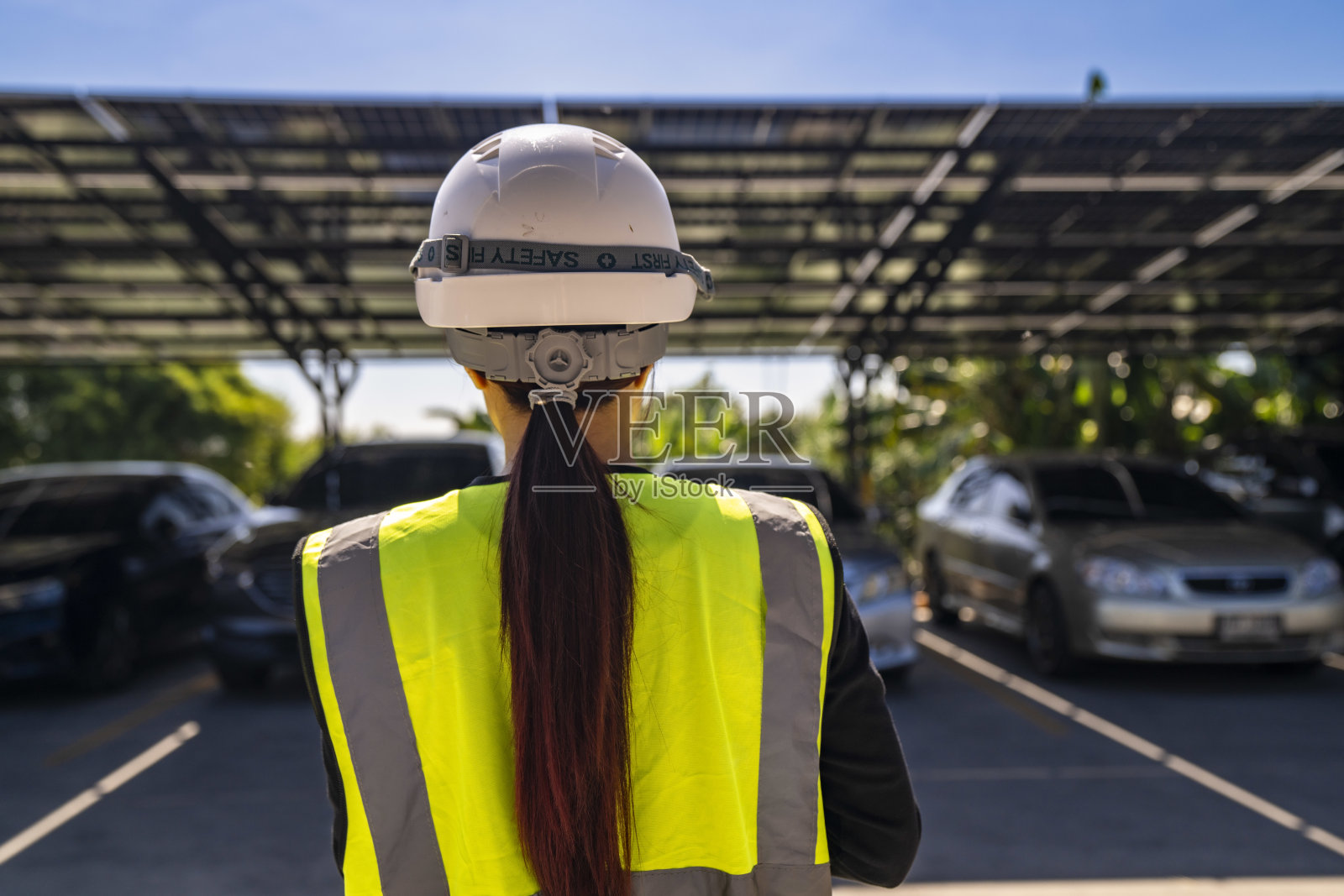 穿着安全装备和头盔的女性工程师正在检查一个太阳能车棚。她观察到停放的车辆被光伏面板遮蔽,促进清洁能源技术和可持续发展。照片摄影图片