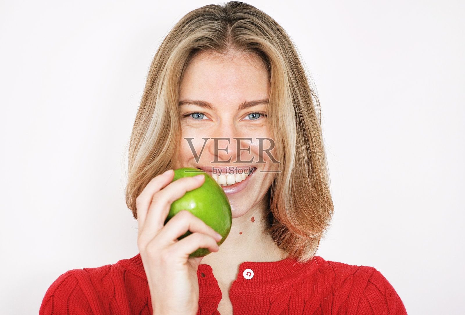 年轻迷人的蓝眼金发短发女孩在浅色中性背景下微笑,手中拿着一个巨大的绿色苹果。照片摄影图片