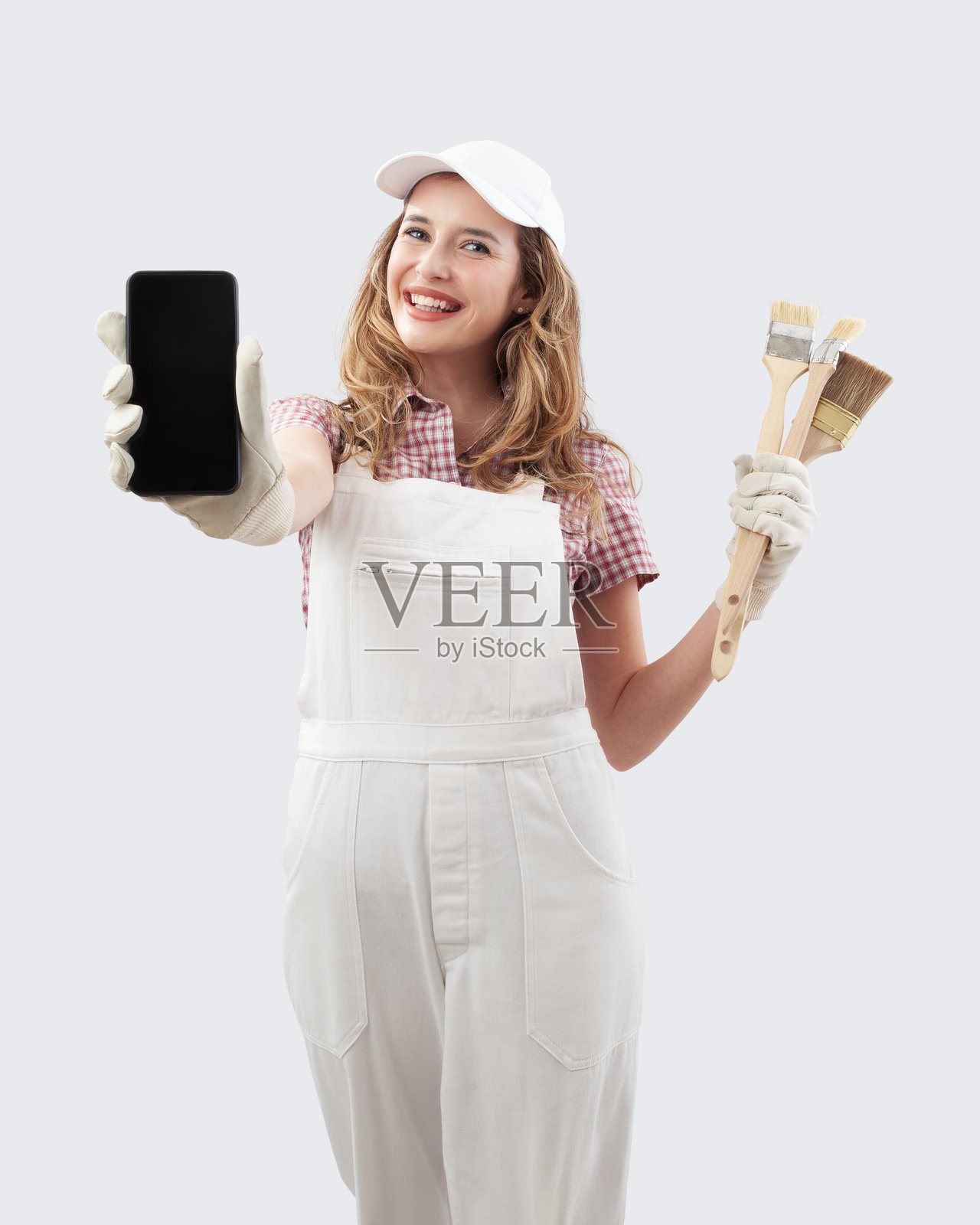 穿着画家工作服的女性向镜头展示手机,进行在线购物以选择墙面涂料和家居装修产品,背景为白色,手中拿着画刷。照片摄影图片