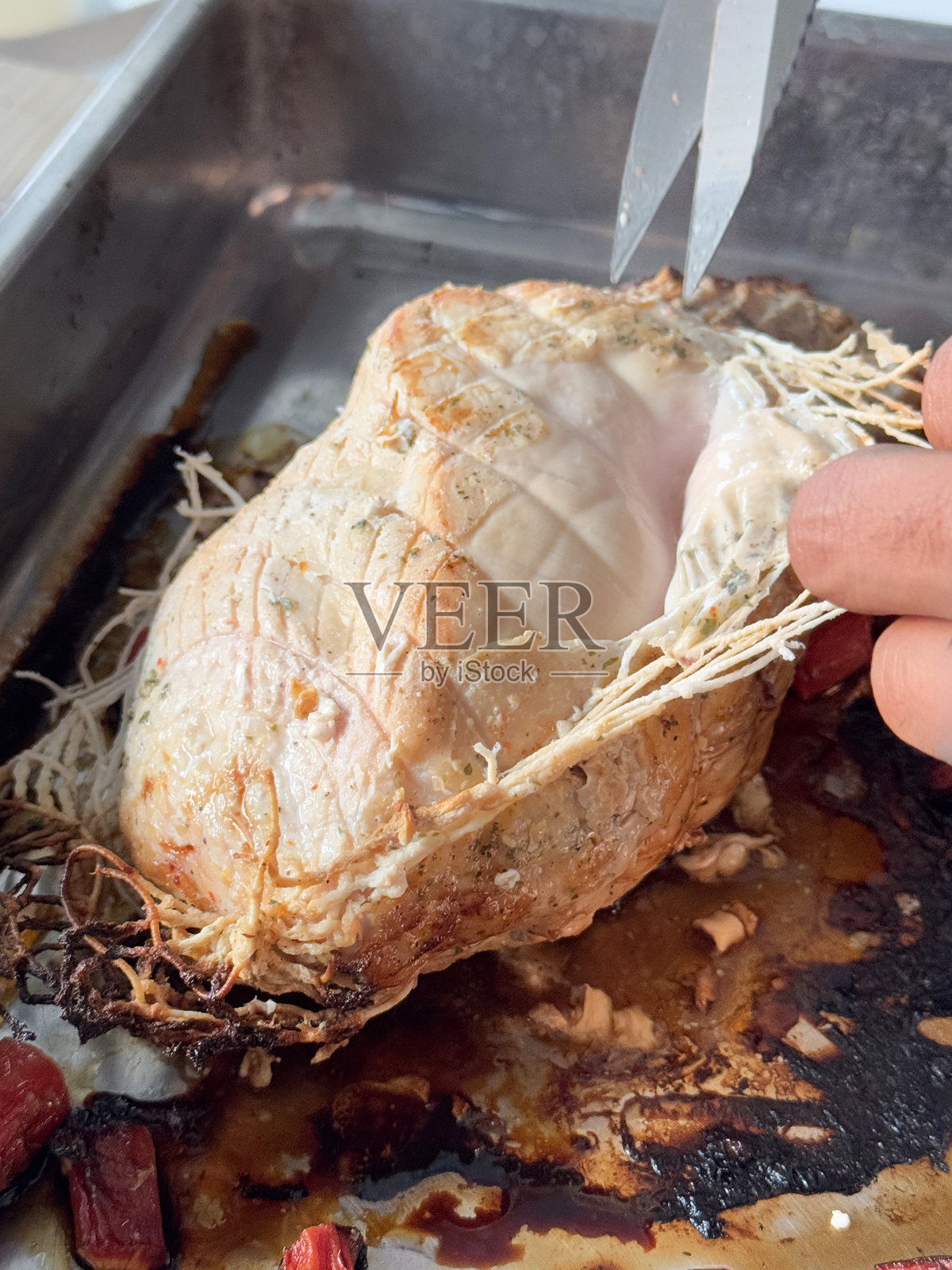 全景图像中,一个无法辨认的人正在用剪刀去除整只烤火鸡胸肉上的支撑网,背景是烤制的胡萝卜和不锈钢烤盘,视角较高,重点在前景。照片摄影图片