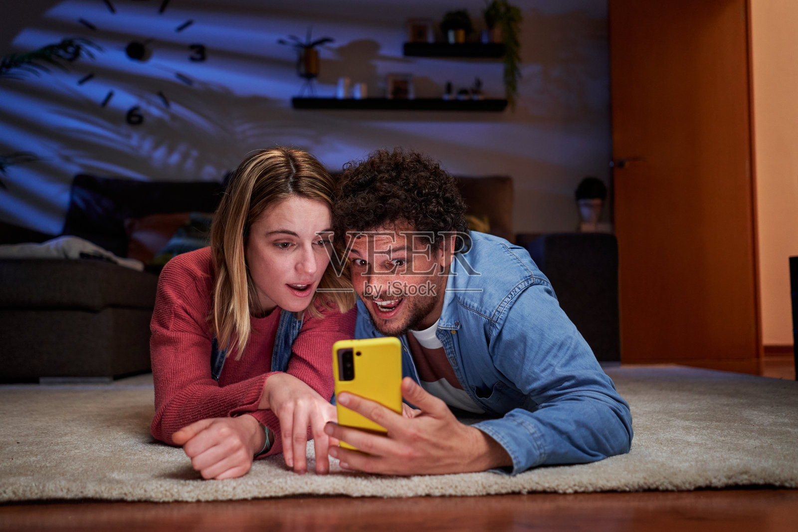 年轻情侣在家夜晚躺在地毯上使用智能手机,表现出惊讶。照片摄影图片