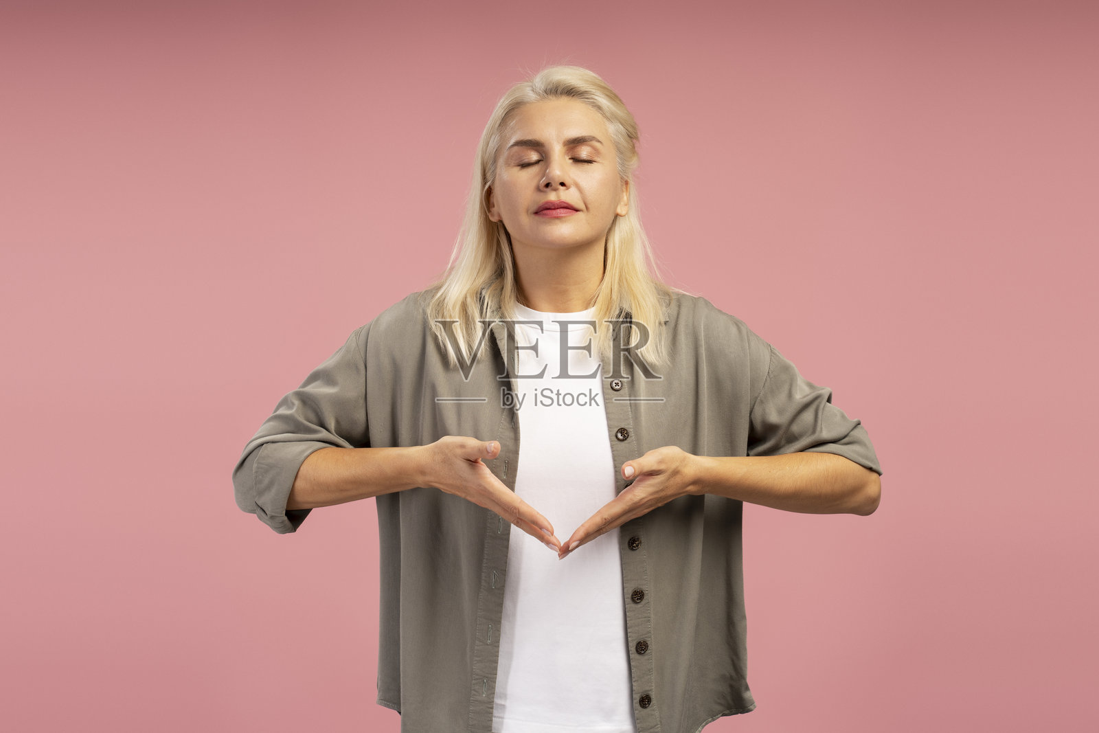 金发成熟的女性在粉色背景下闭眼冥想,双手合十于身体前方。照片摄影图片