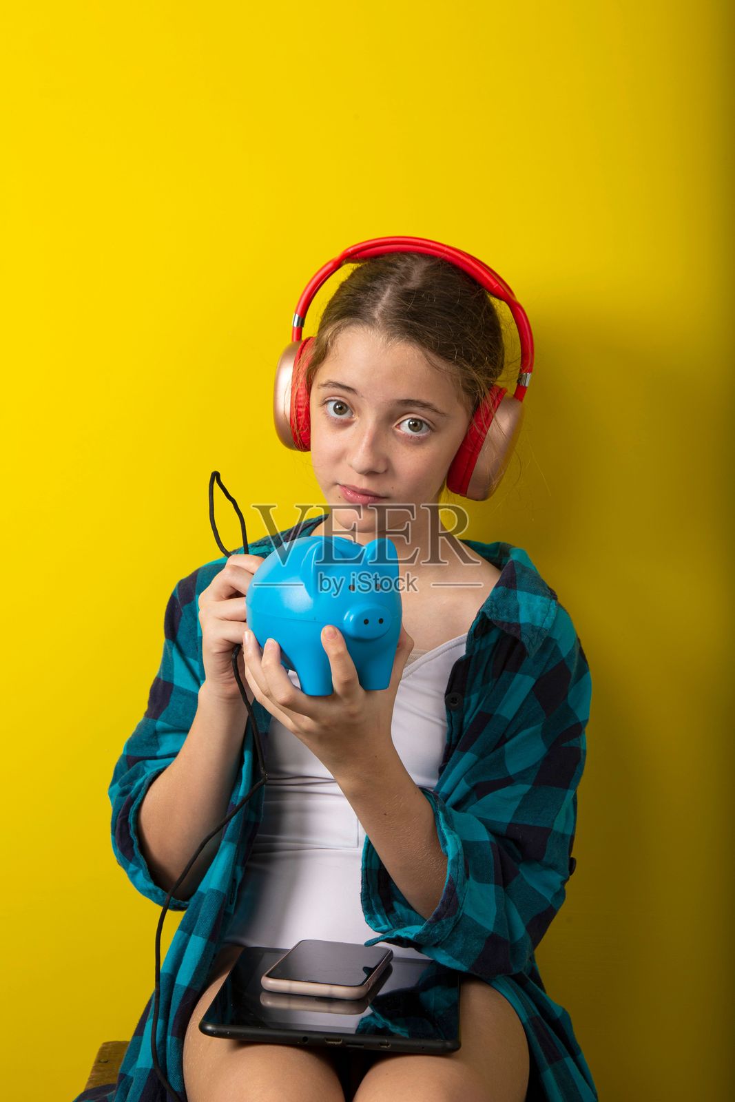 一个戴着耳机的少女在黄色背景下抱着一个蓝色的储蓄罐。照片摄影图片