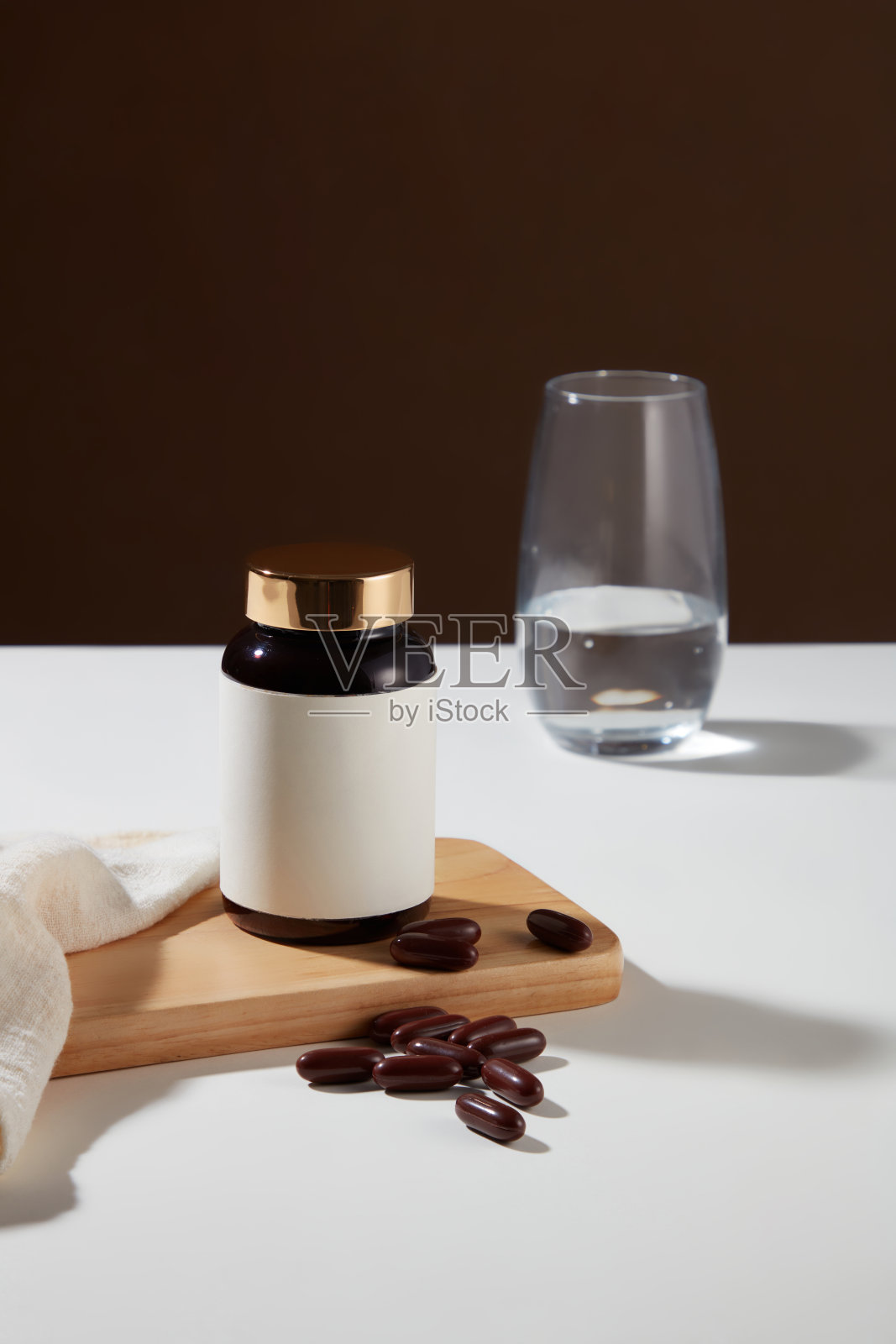 一个没有标签的药瓶放在一个木制平台上,上面放着软胶囊,白色桌子上放着一杯水。深棕色背景。膳食补充剂广告的模型。照片摄影图片