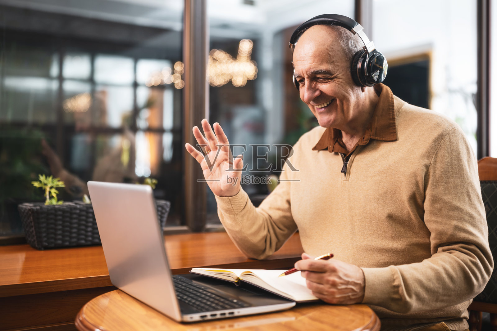 愉快的老年男子在城市酒吧里戴着耳机和笔记本电脑,边学习边挥手。照片摄影图片