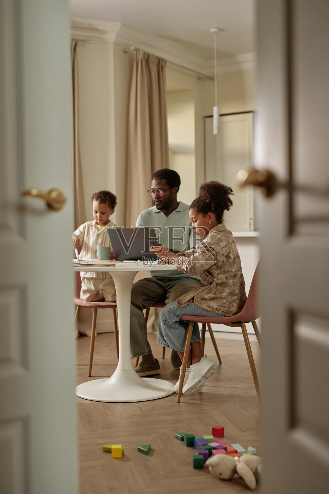 黑女孩在客厅的桌子旁学习,旁边是忙碌的爸爸和小弟弟。照片摄影图片