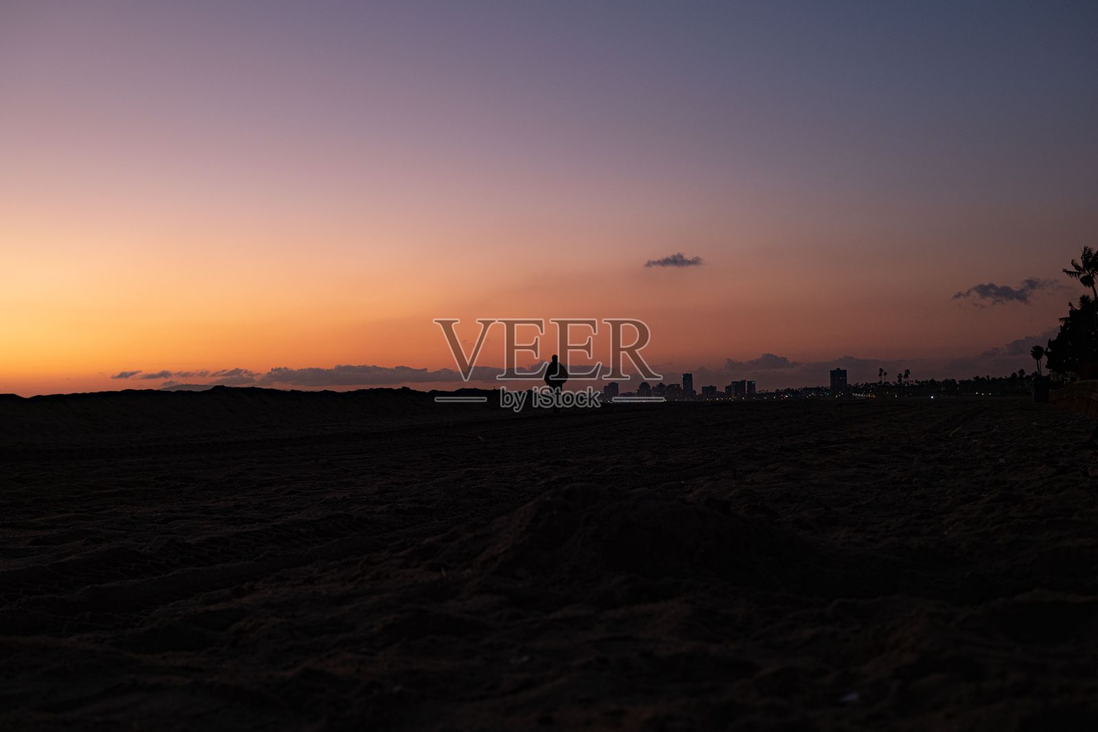 日落时分,海滩上一个人的剪影,背景是城市天际线。照片摄影图片