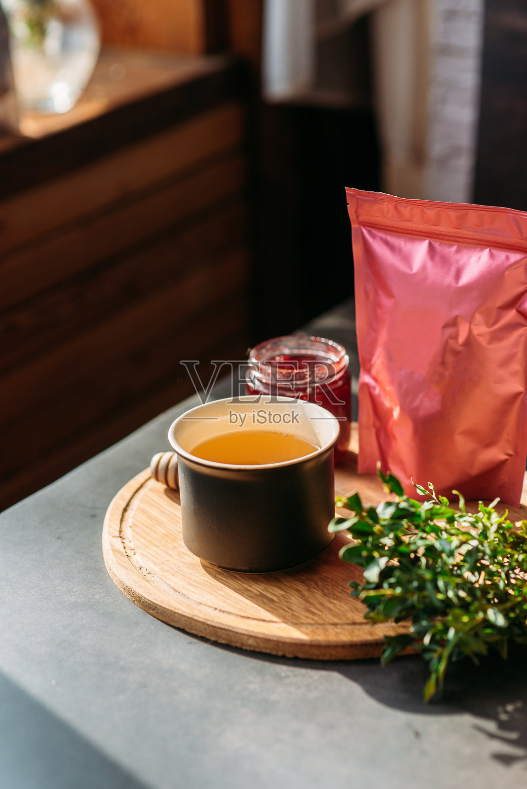 黑色哑光杯中的绿茶,旁边是装有咖啡或茶的拉链包装样品,一个果酱罐,一把木勺盛蜂蜜,阳光透射,早晨的茶,窗边的构图,背景为深色,绿植点缀。照片摄影图片