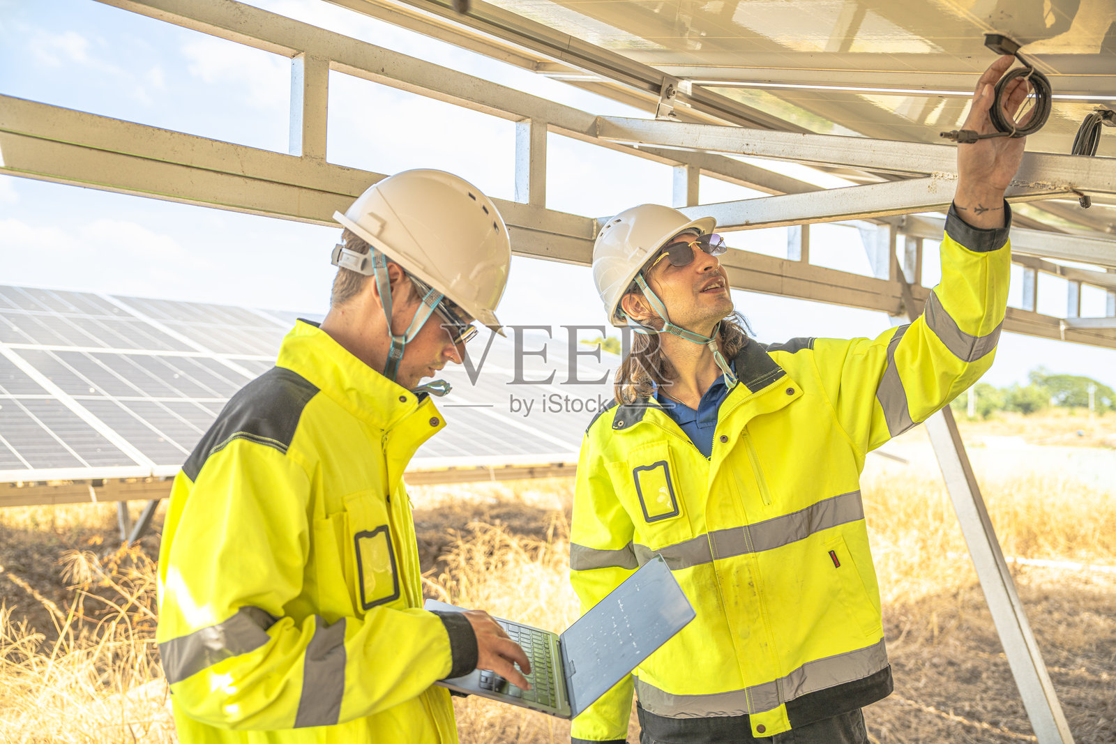 两名穿着亮色反光夹克和安全帽的太阳能技术人员正在检查太阳能板下的电线。一人手持笔记本电脑进行数据记录,另一人则仔细检查电气连接。照片摄影图片