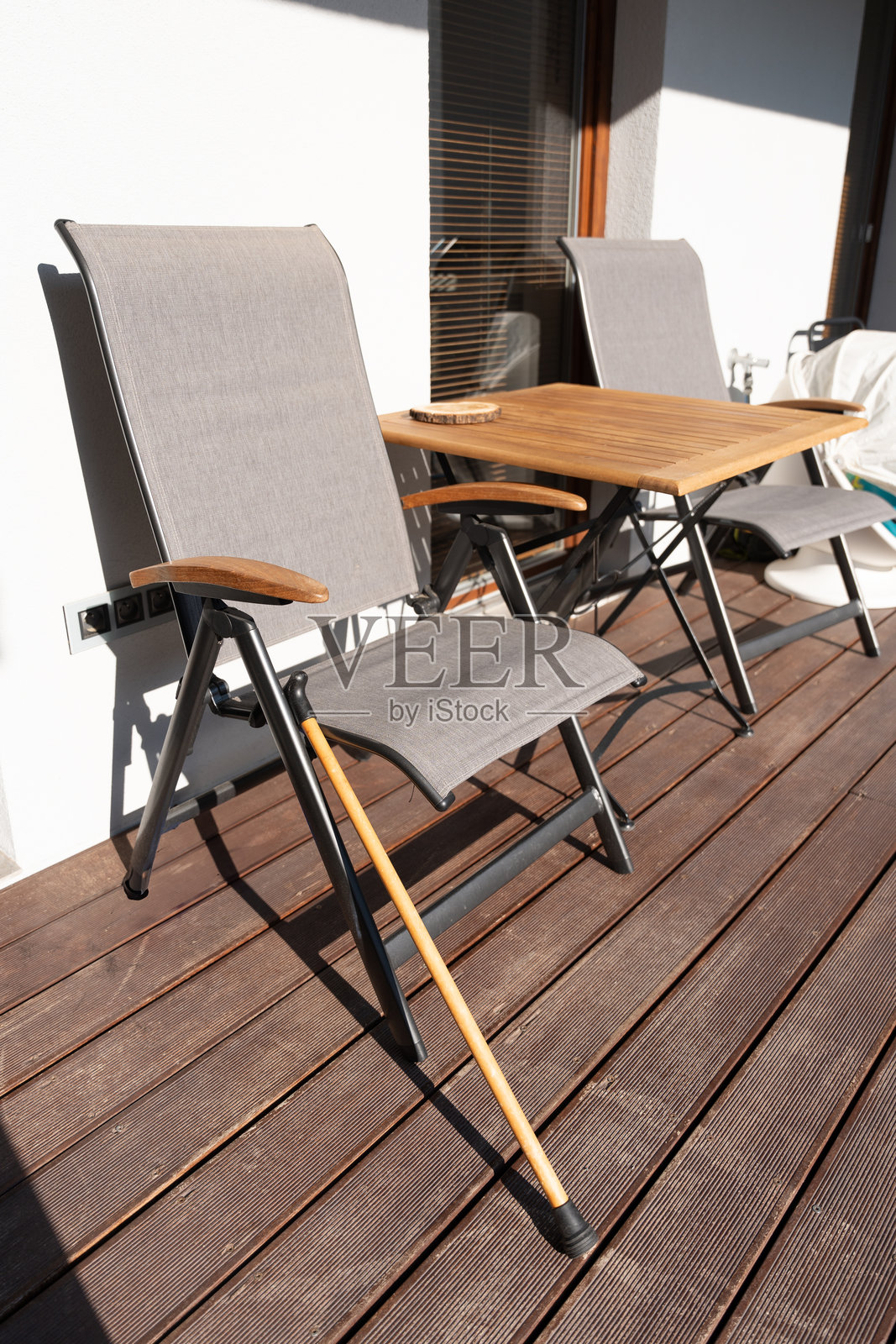 一根手杖靠在现代露台椅子上,阳光洒在木质甲板和木桌上。照片摄影图片
