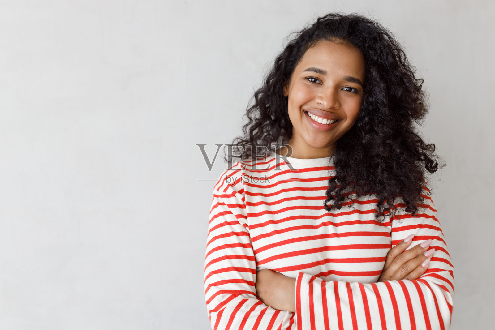 可爱的非洲裔少女穿着红白条纹衬衫,双臂交叉,站在灰色摄影棚背景前,右侧留有空白可用于广告内容或文字。照片摄影图片