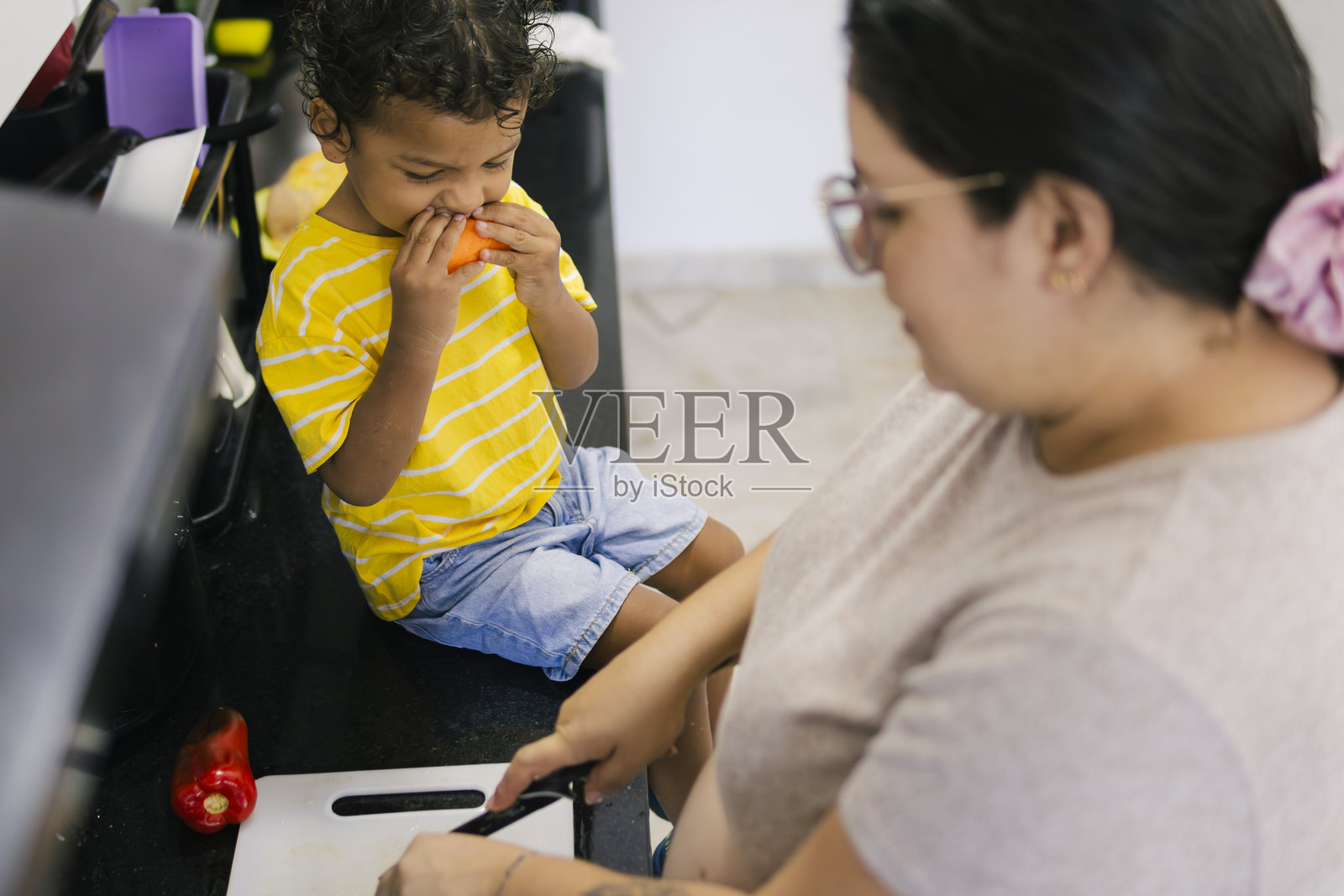 拉丁母亲在厨房里切菜,她的儿子正在吃番茄。照片摄影图片