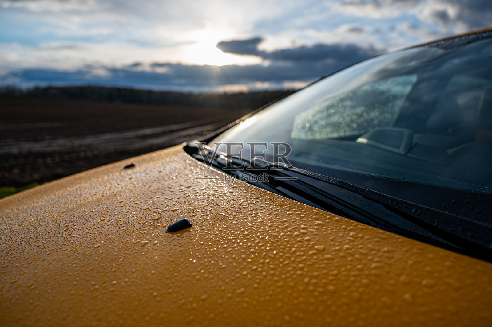 黄色汽车的引擎盖和挡风玻璃上覆盖着水滴,背景是乡村风景和日落时分的戏剧性天空。照片摄影图片