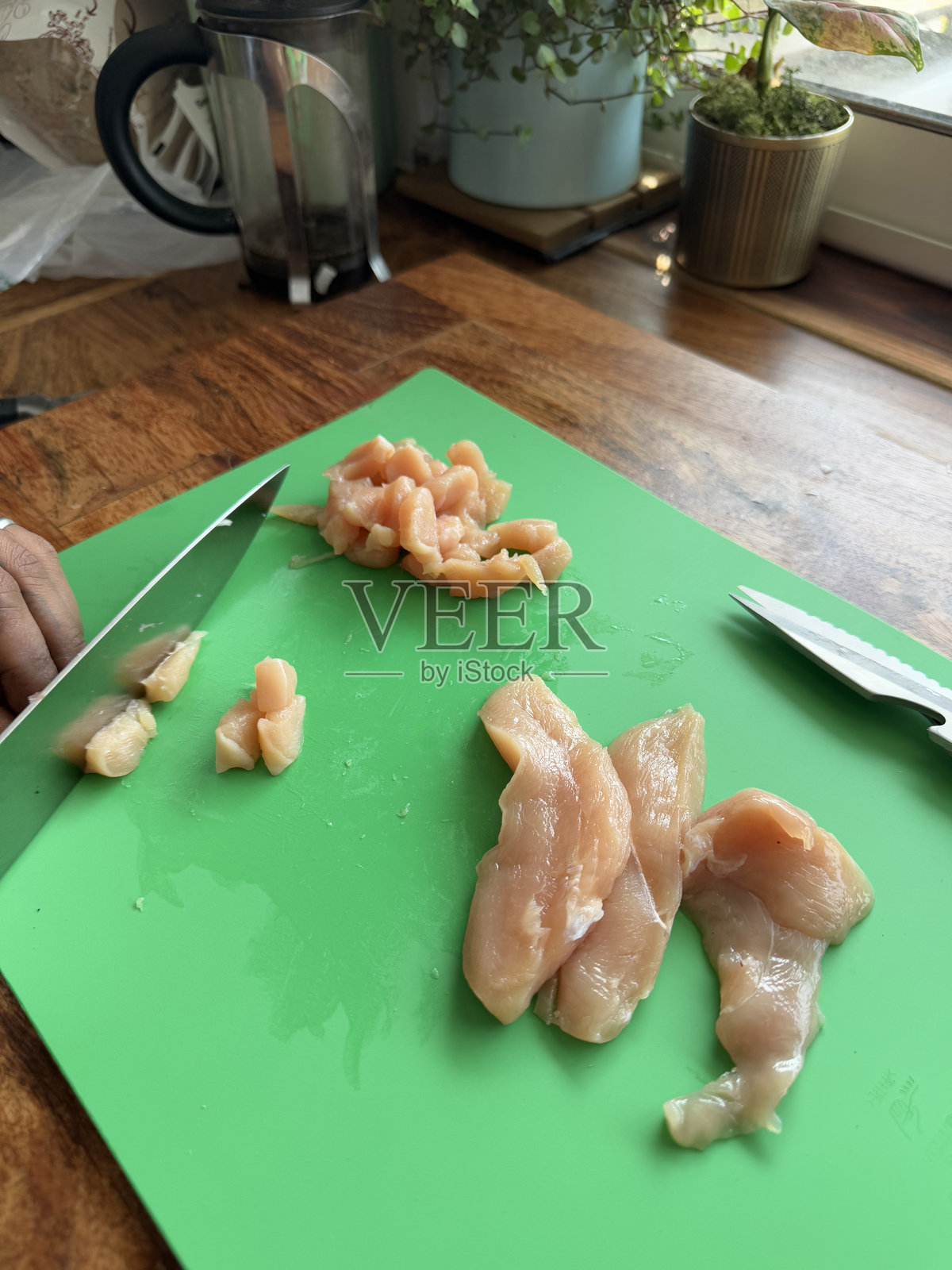 在家庭厨房中,一位无法辨认的人正在塑料砧板上切割生鸡胸肉,男人手持锋利的刀片切割肉类,木质厨房台面,俯视角度,重点在前景。照片摄影图片