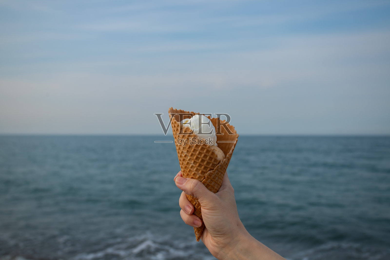 手握冰淇淋甜筒,海洋背景下的海滩美食,阳光下的冰淇淋在蓝色水面旁。照片摄影图片