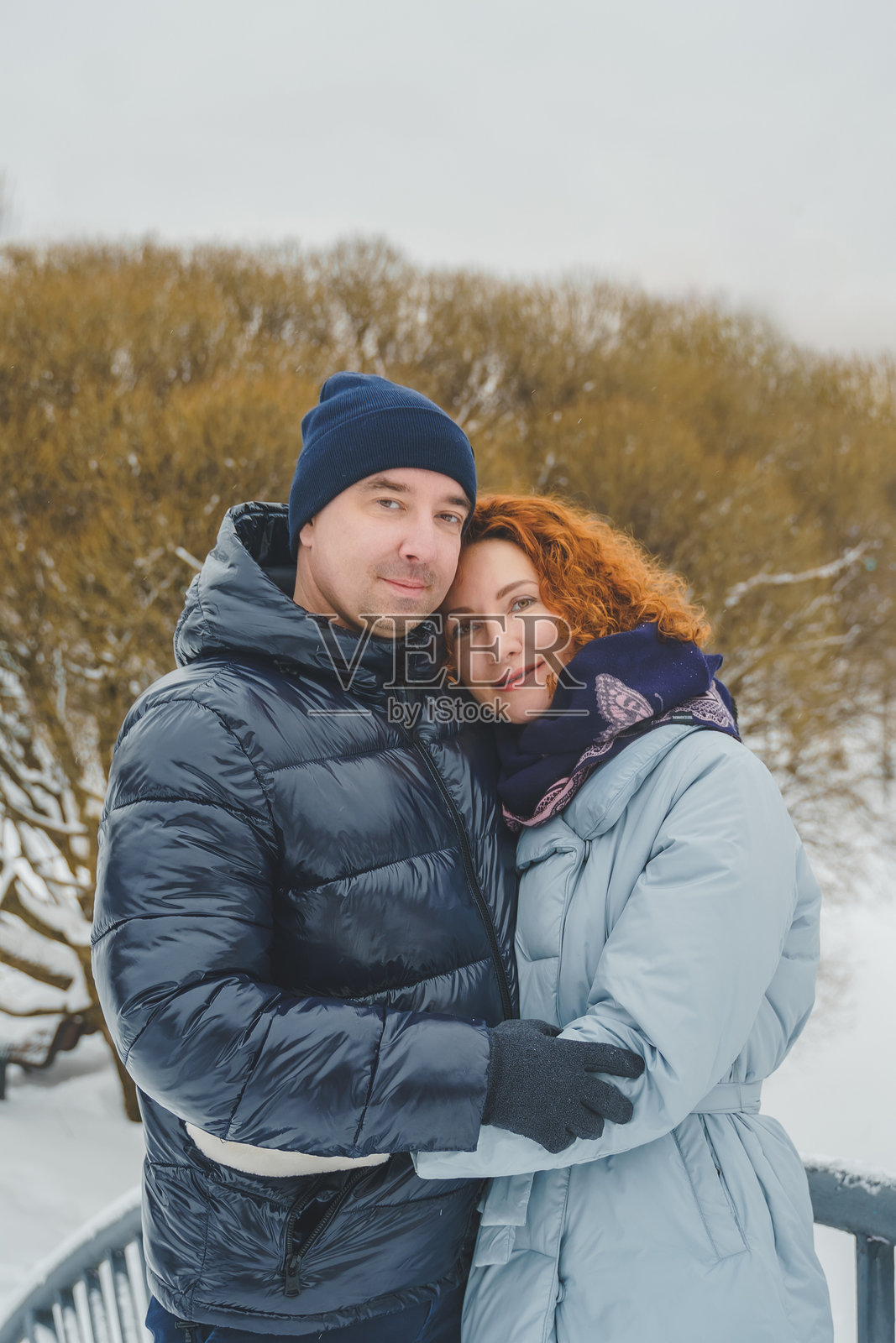 浪漫爱情故事。快乐的中年千禧夫妇,冬季雪地公园里一位红发女子。浪漫约会,亲吻,拥抱。散步,享受乐趣。时尚的蓝色衣服,夹克,帽子。照片摄影图片