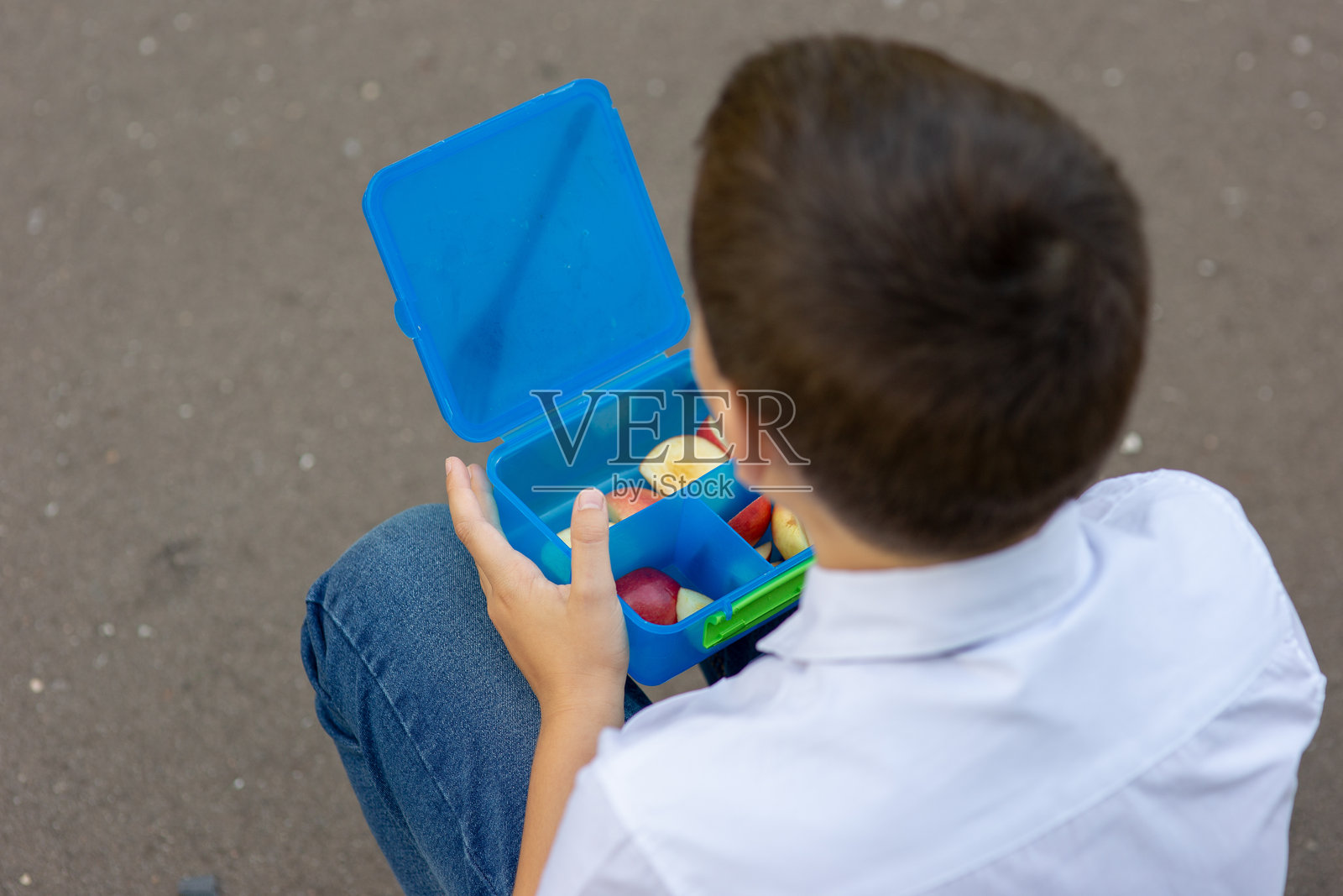 穿着白衬衫的男孩背对着镜头,手里拿着装有苹果的蓝色午餐盒。照片摄影图片