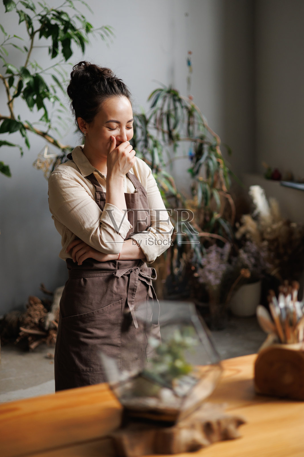 快乐的亚洲花店老板穿着围裙,在模糊的植物园和工具旁边笑着照片摄影图片