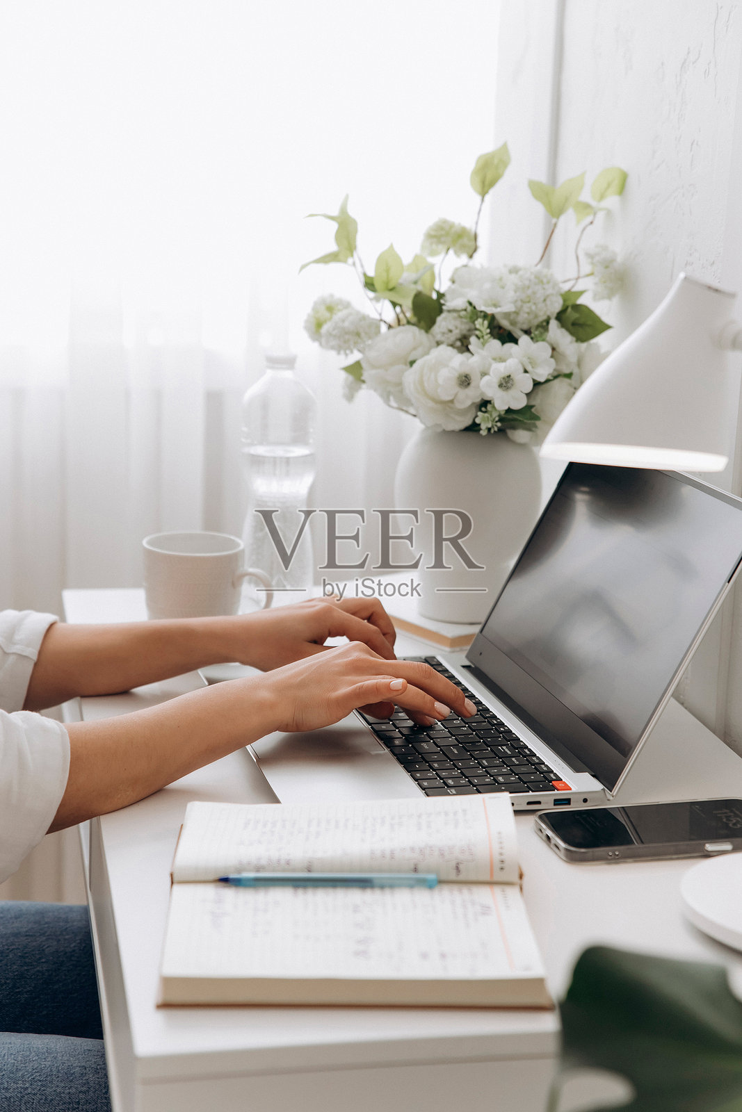 在白色的桌子上,女人的手在笔记本电脑键盘上打字的特写,背景是一瓶饮用水和花瓶里的花。自由职业的概念,远程工作,在线学习照片摄影图片