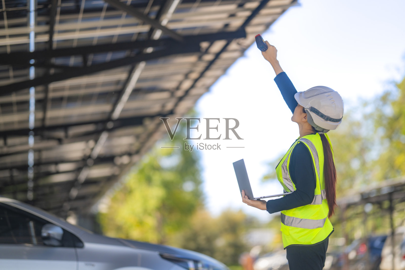 一名女性技术人员戴着安全头盔和反光背心,使用笔记本电脑检查太阳能车棚的安装。场景中有太阳能电池板和停放的汽车,天空晴朗。照片摄影图片