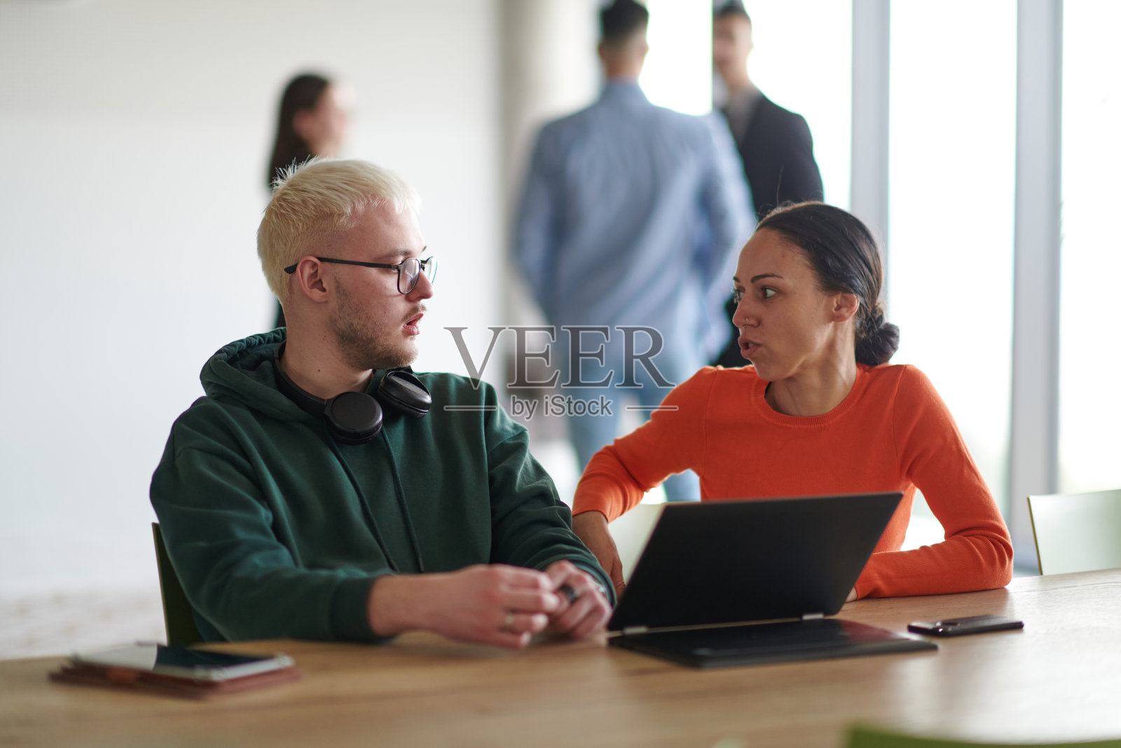 一位金发商人与他的同事坐在办公室里讨论工作,同时使用笔记本电脑协作完成任务。照片摄影图片