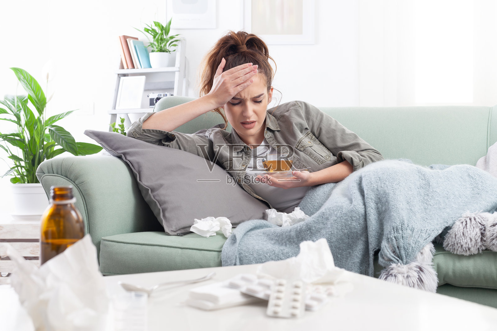 年轻女性坐在沙发上休息,捂着头感到疼痛。她身体不适,正遭受头痛,手中拿着一杯茶。前景中有纸巾、糖浆瓶和药丸,展现了生病与康复的场景。照片摄影图片