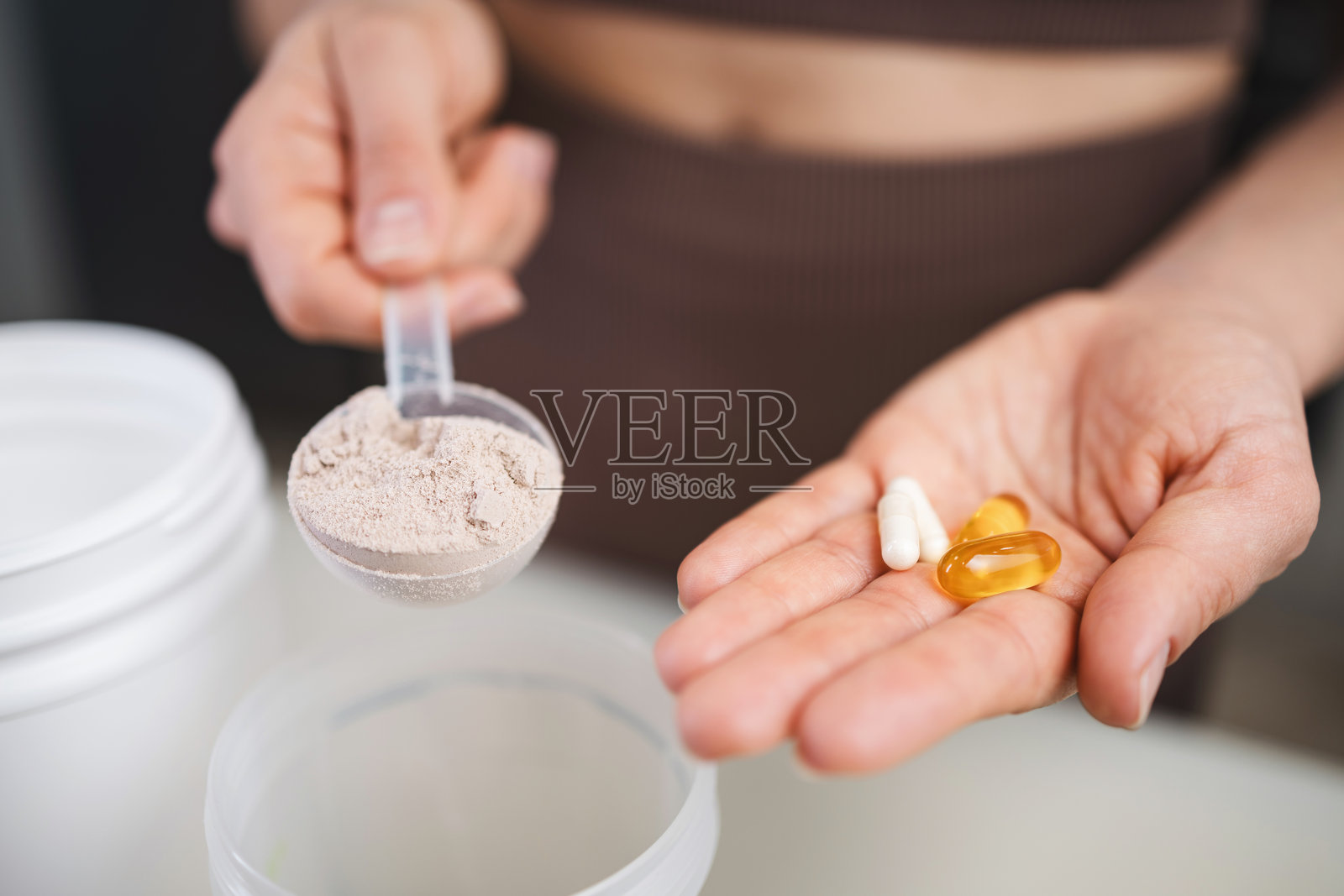 穿着运动服的运动女性在厨房准备蛋白饮料,手中拿着一把量勺,里面装有乳清蛋白粉和Omega-3补充剂,另一只手握着白色氨基酸胶囊或维生素。照片摄影图片