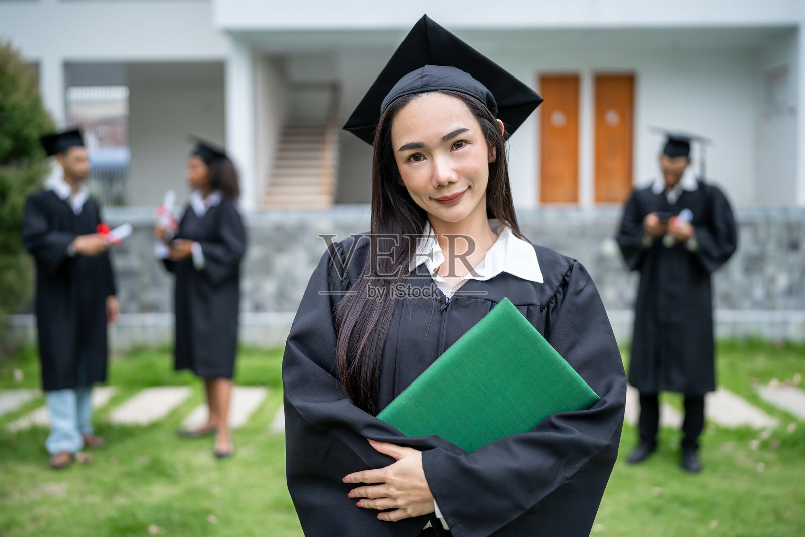 庆祝大学成就的亚洲女研究生肖像。兴奋的迷人成功女孩穿着毕业袍和学位帽,站在大学外面看着镜头。照片摄影图片