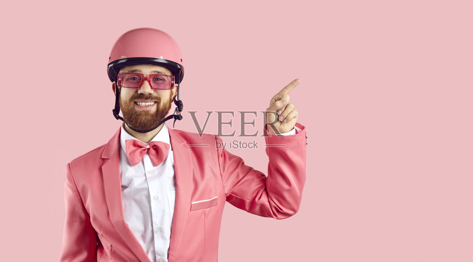 穿着西装、蝴蝶结和眼镜,戴着粉色自行车头盔的搞笑男子微笑着并指向一侧照片摄影图片