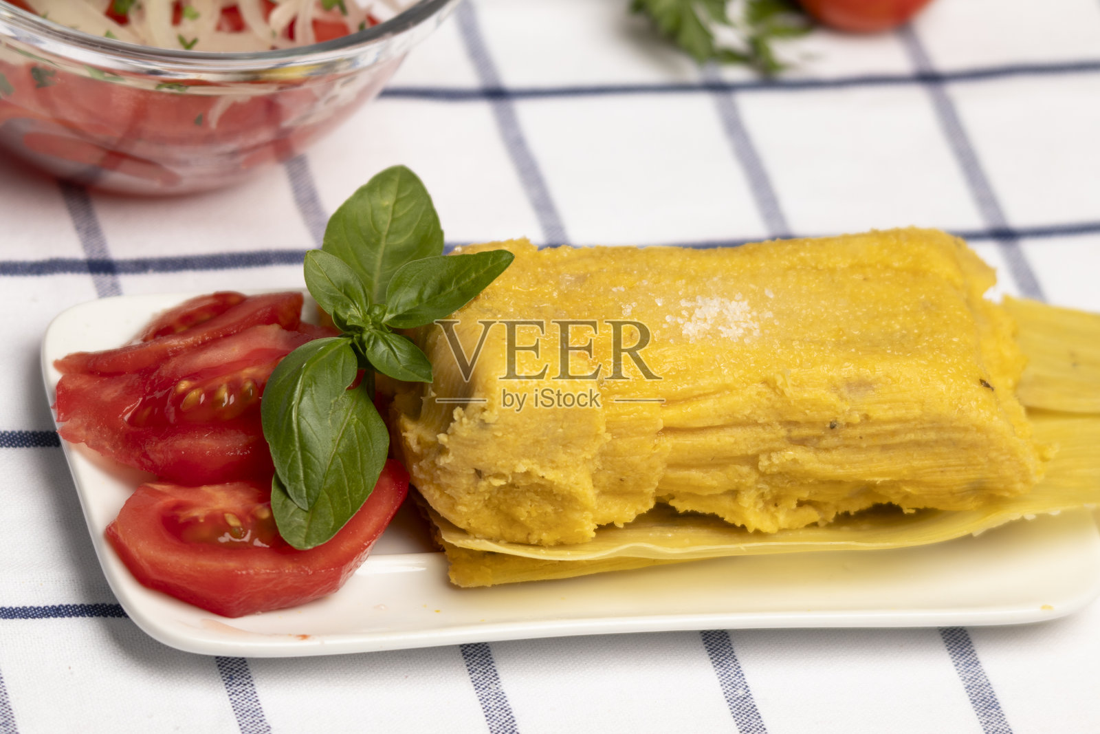 用叶子包裹的玉米饼,配有番茄和罗勒叶,典型的智利夏季菜肴,背景是一碗盛有洋葱和新鲜成熟番茄的番茄沙拉。照片摄影图片