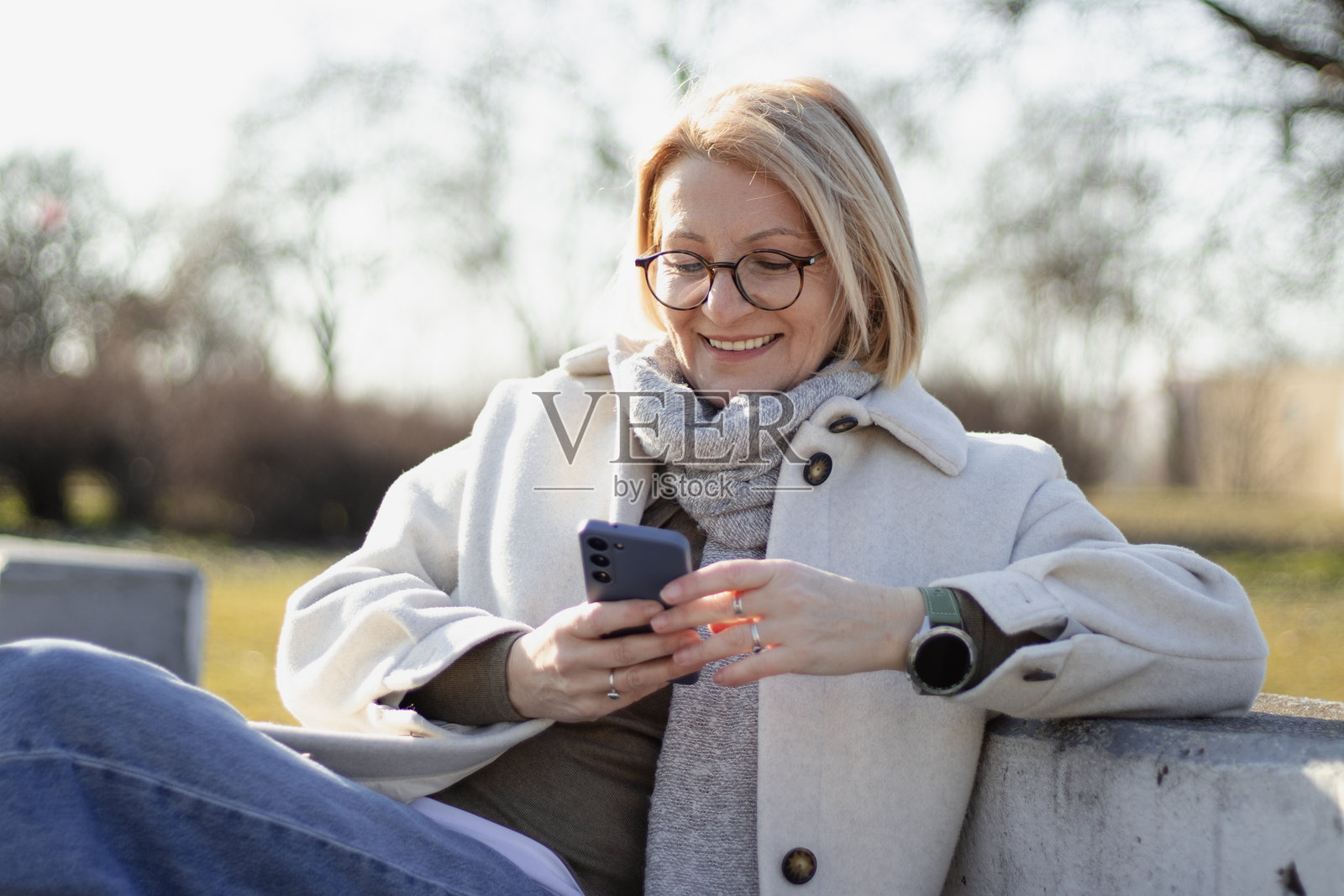 成熟的金发快乐女性戴着眼镜,坐在公园长椅上,在阳光明媚的日子里一边享受大自然,一边使用手机。照片摄影图片