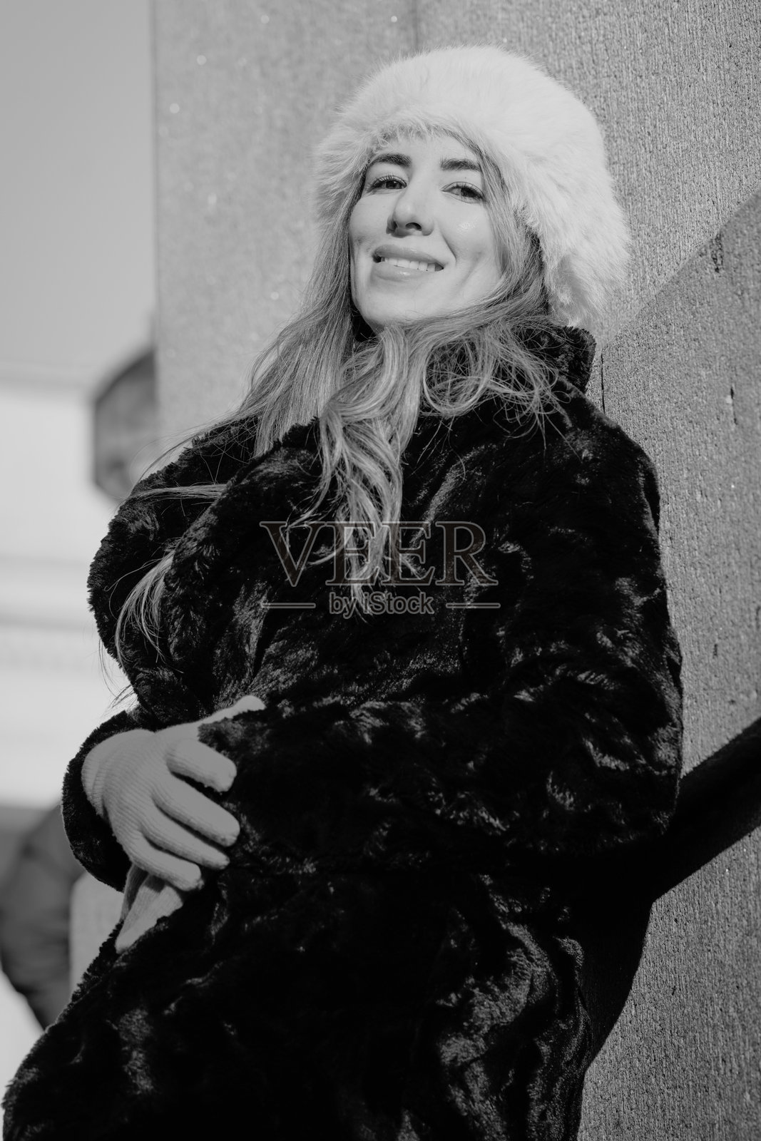 金发女性模特穿着黑色仿毛皮外套,倚靠在石墙上 - 户外中等肖像照,注视着镜头照片摄影图片