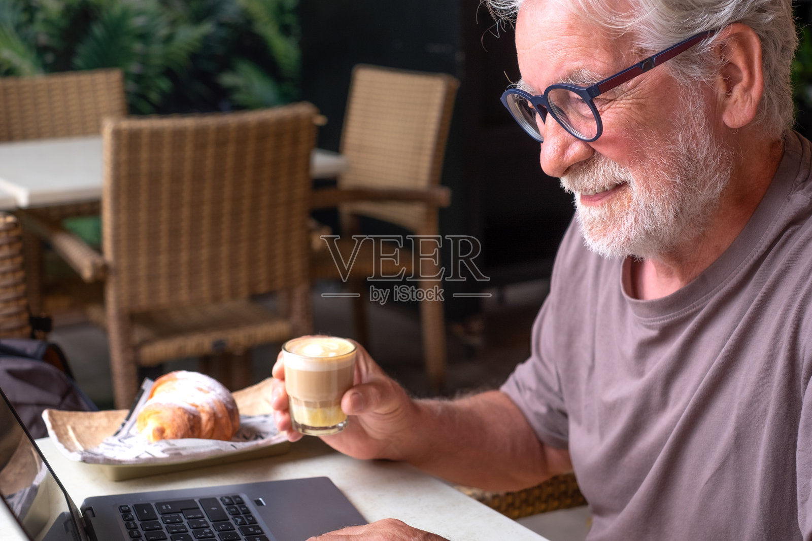 一位帅气的老年男性在咖啡馆享用早餐,手边有咖啡和可颂,正在使用笔记本电脑。他微笑着,放松地在咖啡馆的氛围中敲击键盘。照片摄影图片