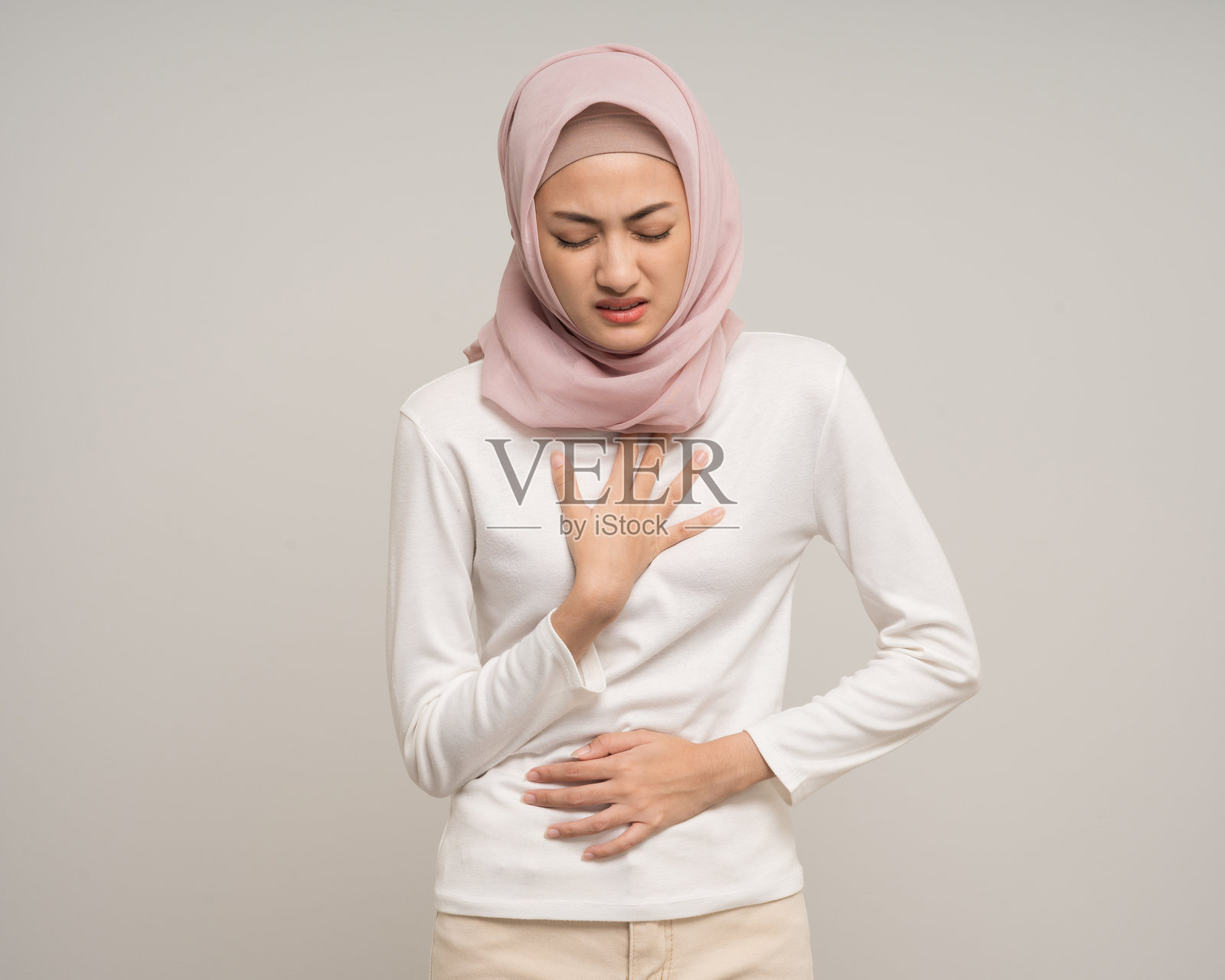 不健康的年轻亚洲穆斯林女性喉咙刺激、黏液和咳嗽。发烧、头痛、呼吸道感染,站在孤立的白色背景上。穆斯林女性穿着阿巴亚和头巾。照片摄影图片