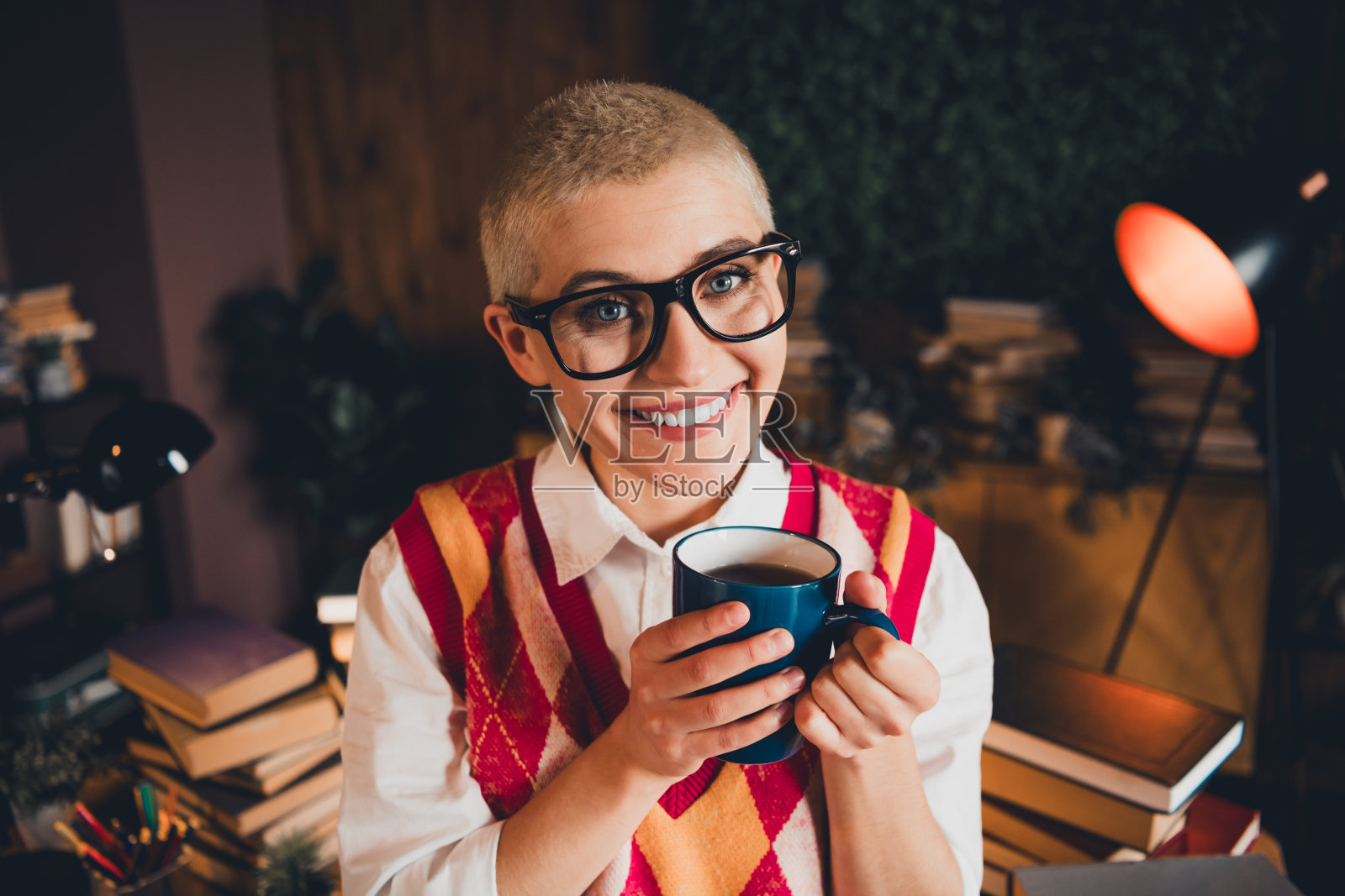 年轻女子穿着色彩斑斓的背心和眼镜,在温馨的学习区享受咖啡,周围是书籍,正值傍晚时分。照片摄影图片