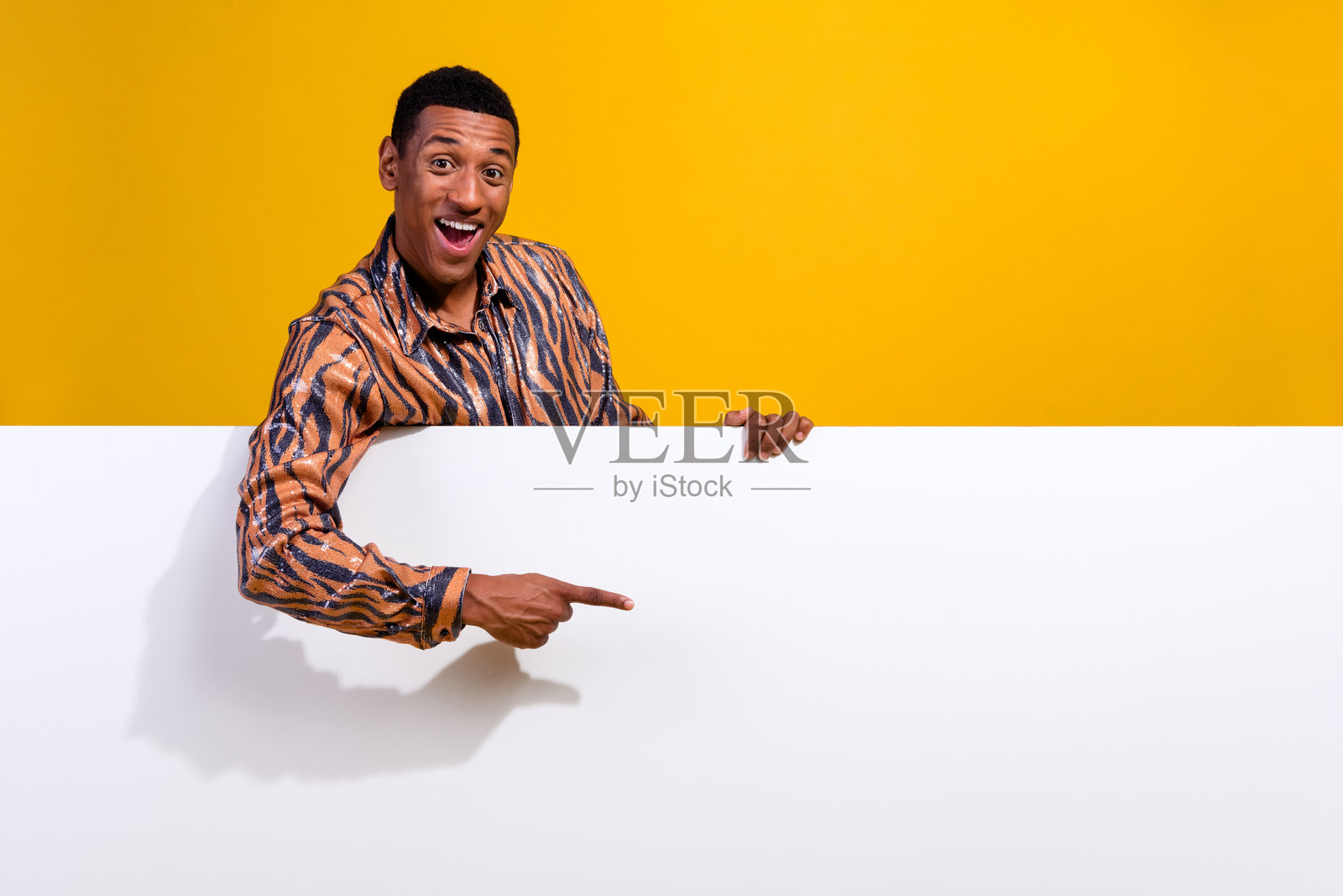 微笑的年轻男子指着黄色背景的空白广告牌,穿着时尚印花衬衫,展示广告概念照片摄影图片